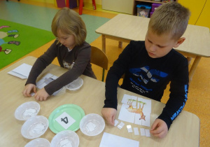 Dwoje dzieci siedzi przy stole na którym mają ułożone obrazki z części. Dzieci układają małe białe papierowe prostokąciki zgodnie z liczbą głosek zawartych w nazwach zwierząt z puzzli.